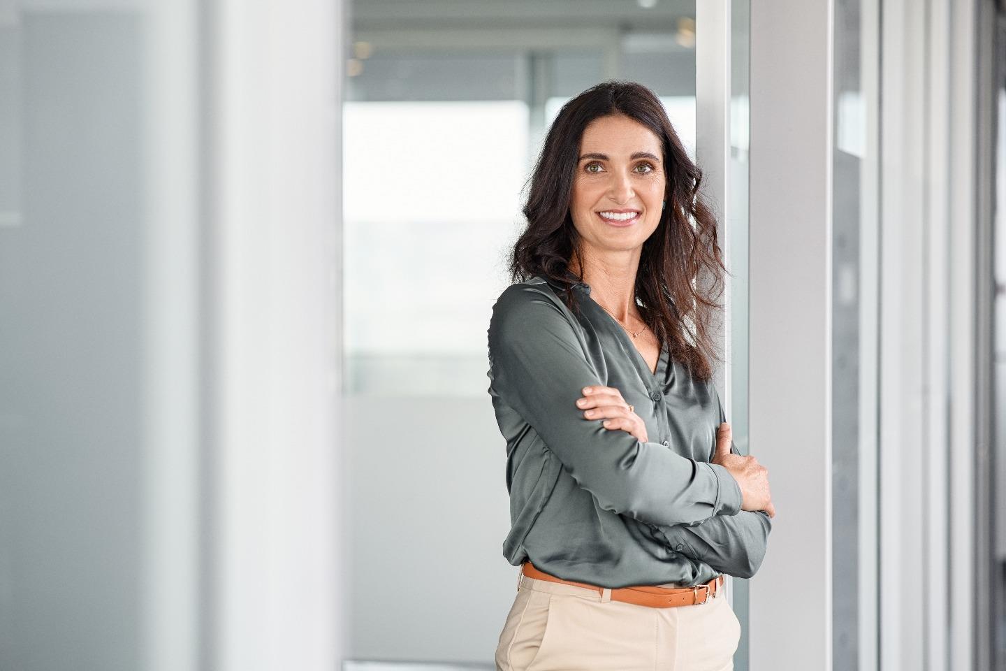 Woman with long hair standing with her body facing the window, her arms crossed and looking into the camera with a smile.